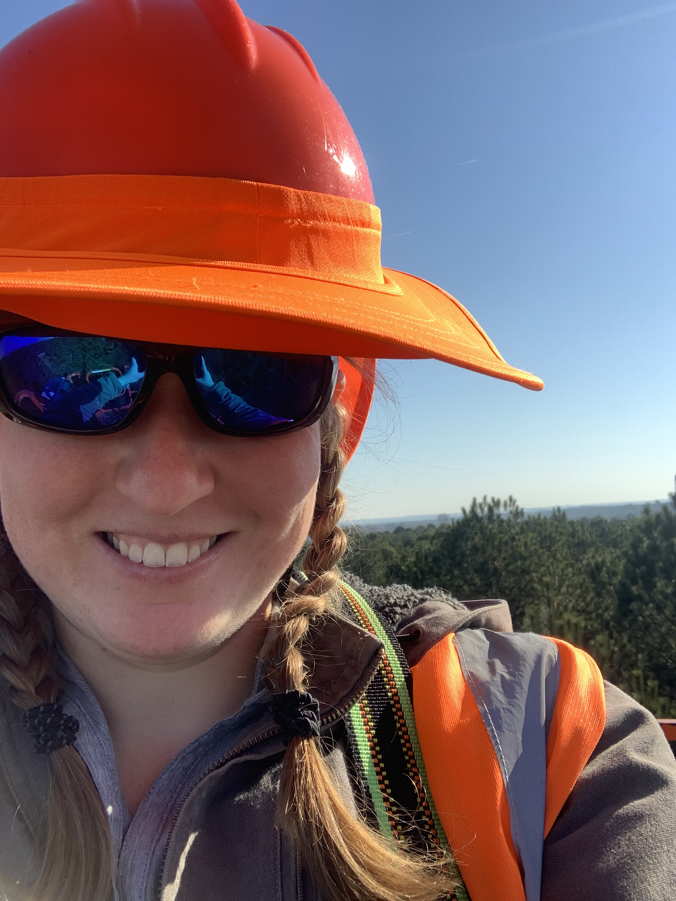 Selfie image of Jaine in a lift. Jaine is wearinga n orange hardhat with a visor attached and sunglasses. The background is a blue sky and the tops of many trees.