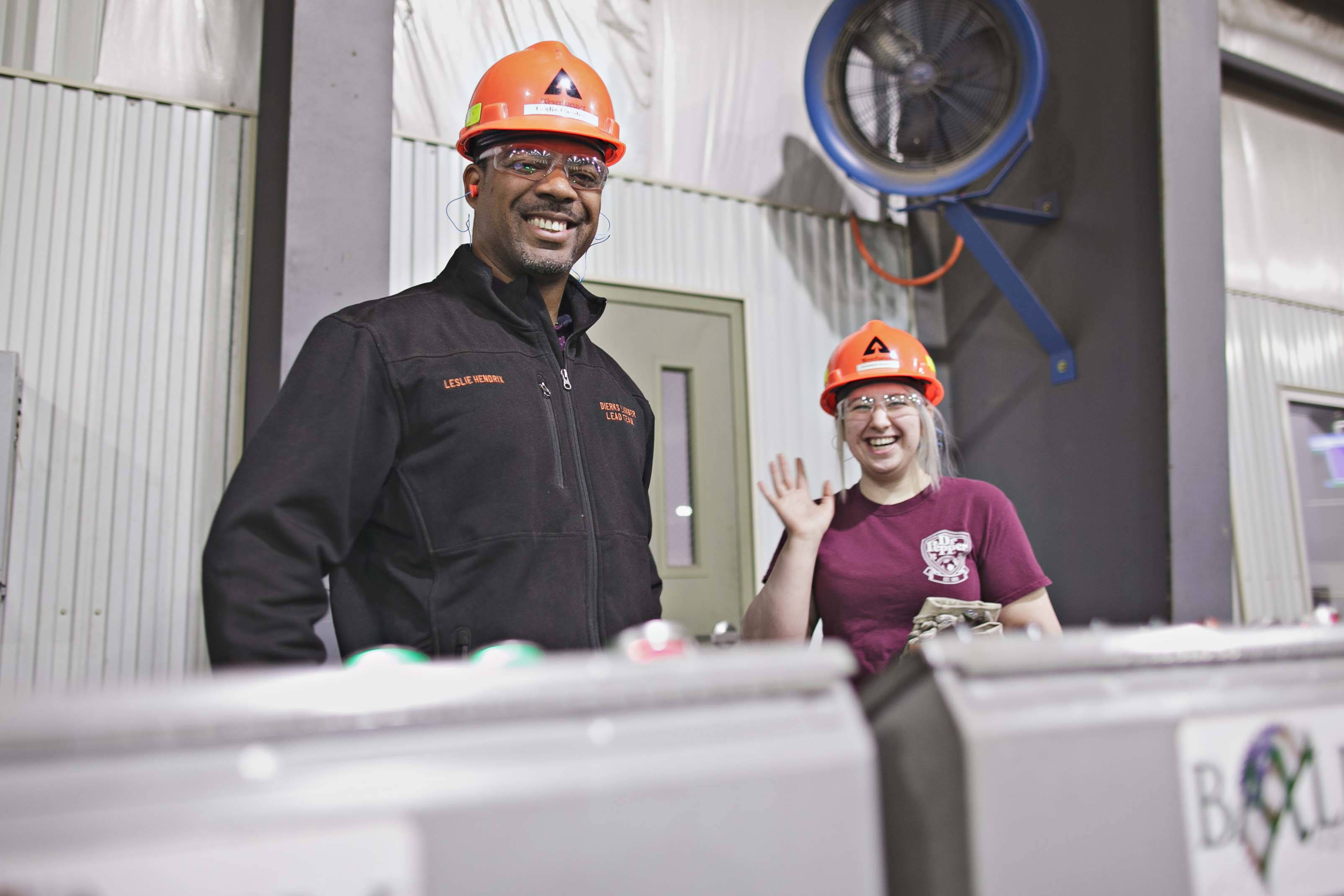 A pair of people wearing hardhats pose. The man, standing on the left, smiles while wearing a hardhat. The woman, to the right, is smiling and waving as she wears a hardhat.