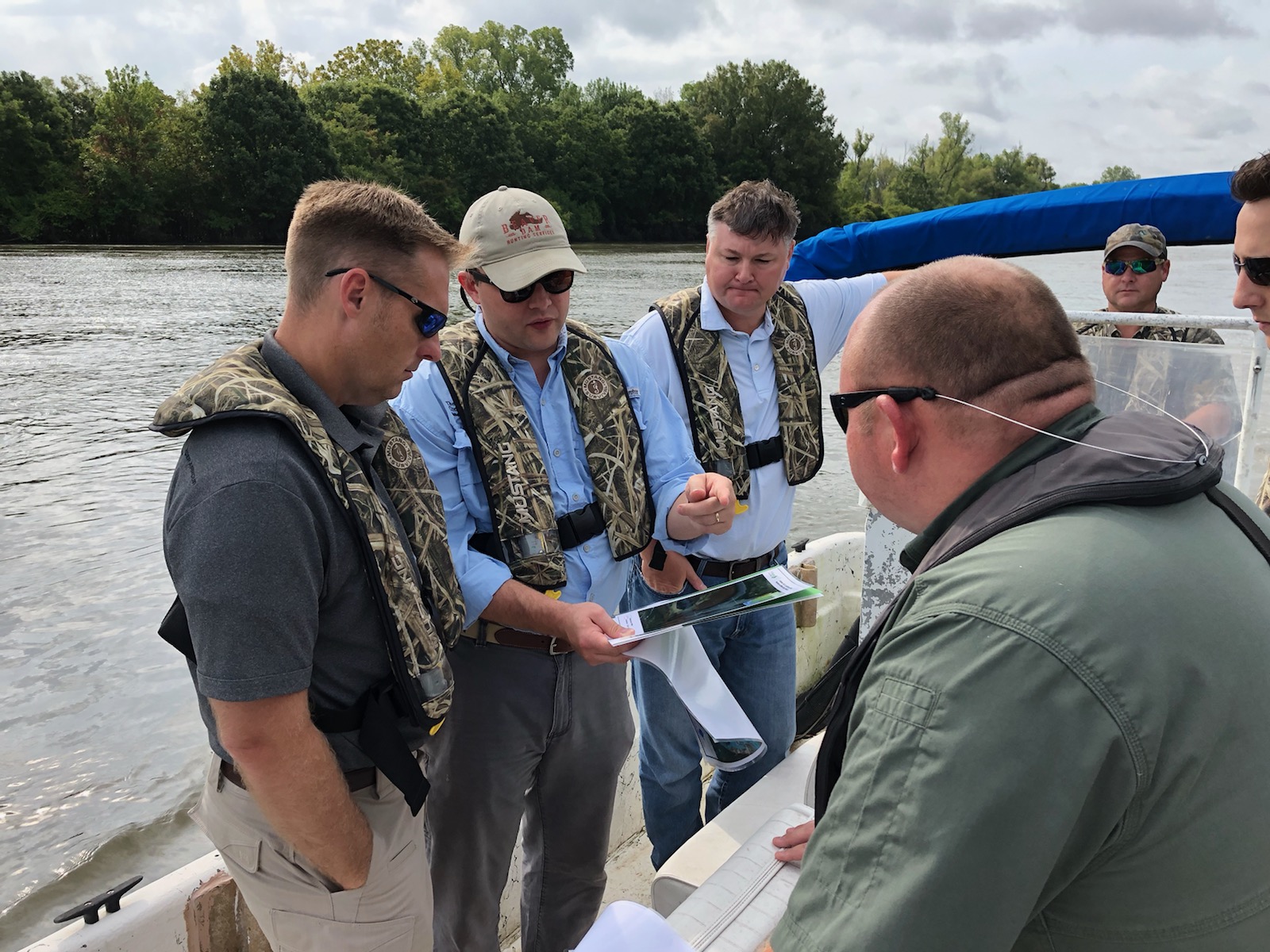 Image of Alex Littlejohn discussing the benefits of flood control management with congressional leaders while he was at the Nature Conservancy. Alex is in the foreground wearing a lifevest and aboard a boat. He is surrounded by other men.