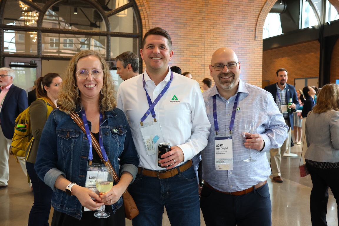 Image of Laura Six, wearing a denim jacket over a dark dress, Taylor Scherer, wearing a white long-sleeve shirt with a Weyerhaeuser logo on the left breast bocket, and Adam Campbell, wearing a blue button-down shirt, in the lobby at the conference.