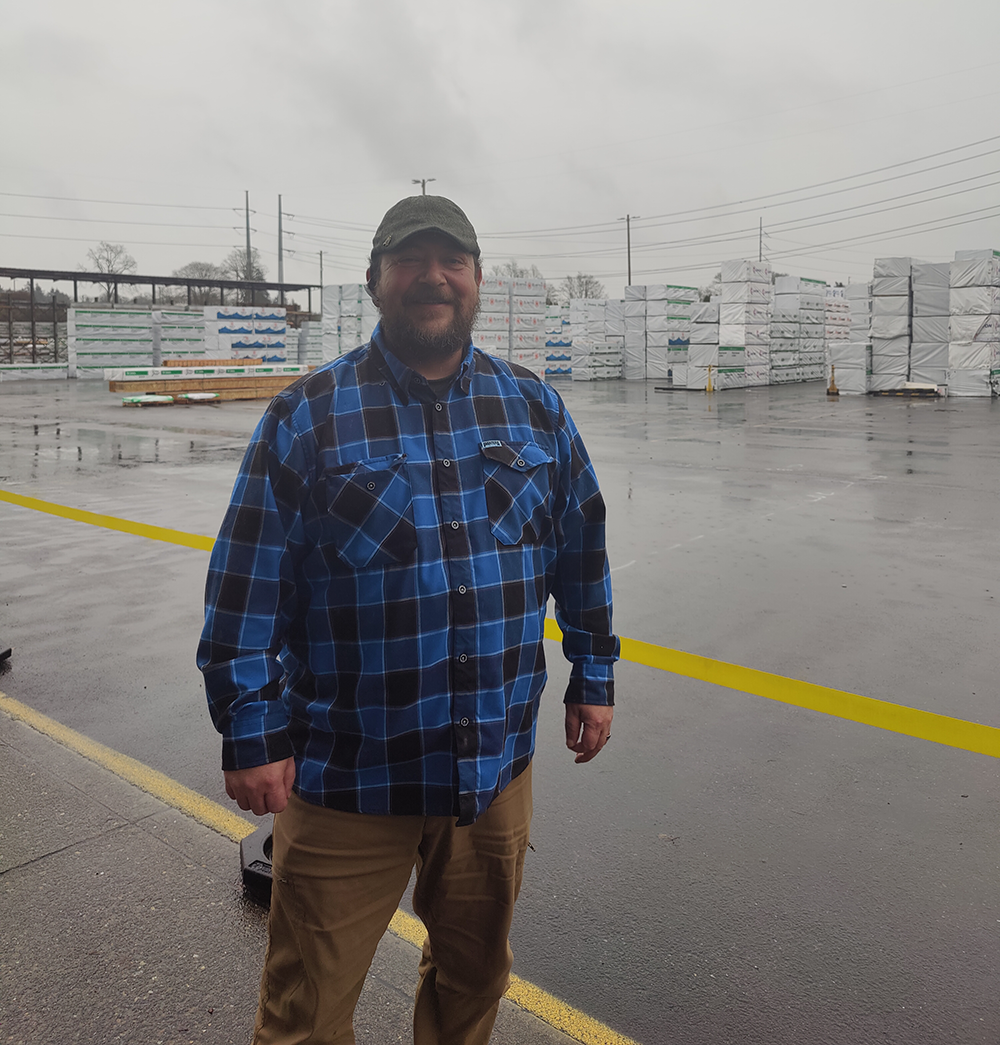 Image of Bob Jewell at the Tacoma distribution center on a rainy day. Bob is wearing a jeff cap and blue flannel shirt with kahki pants. He is smiling as timber products in Weyerhaueser wrappers are in the background.