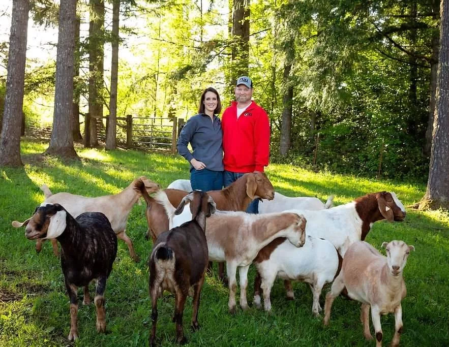 Image of Jill standing with her husband while surrounded by goats. They are standing outsdie on green grass with trees in the background.