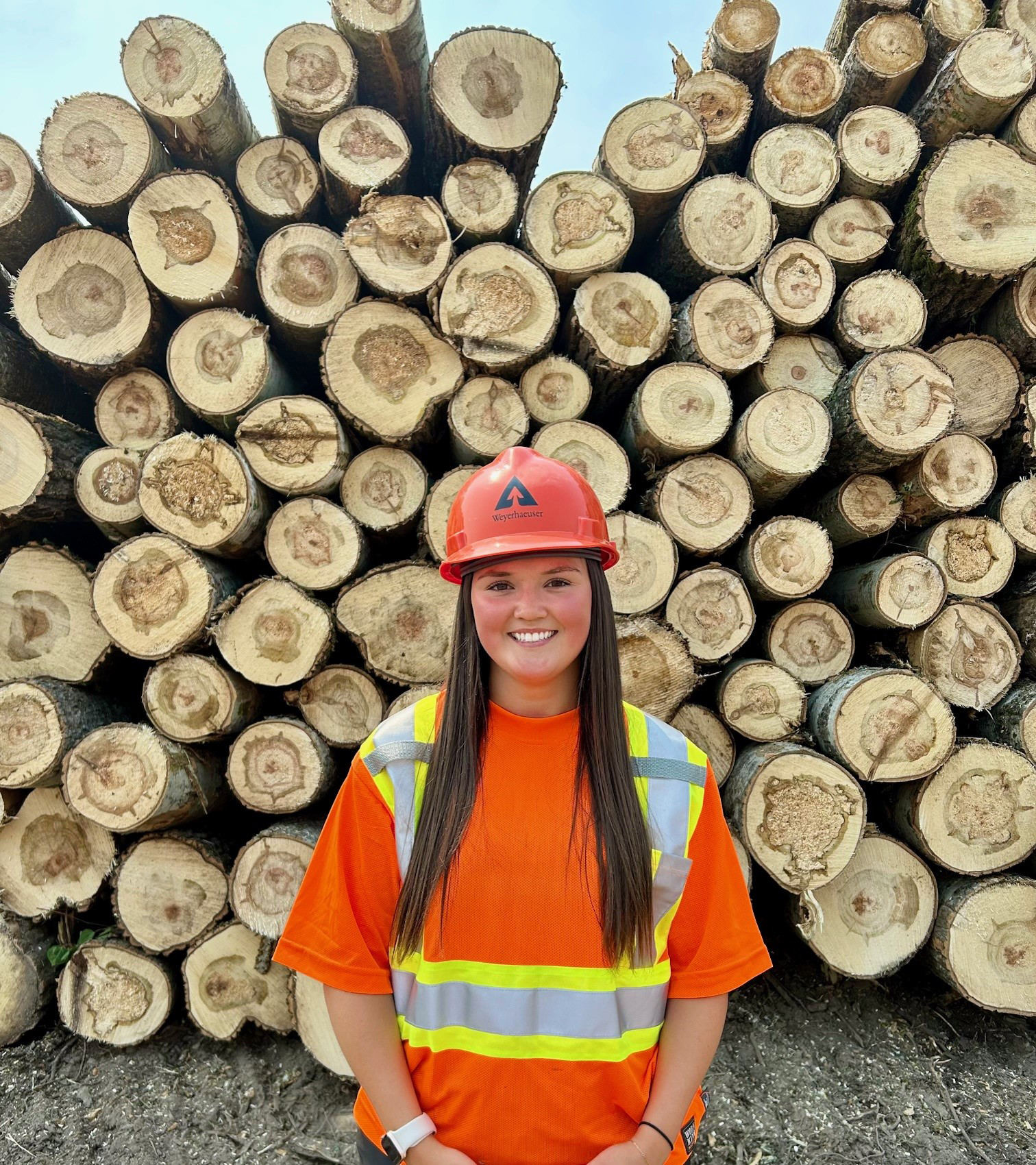 Image of Logan standing in front of a stack of freshly cut logs. Logan is wearing personal protective gear with an orange helmet, orange safety shirt and yellow safety vest.