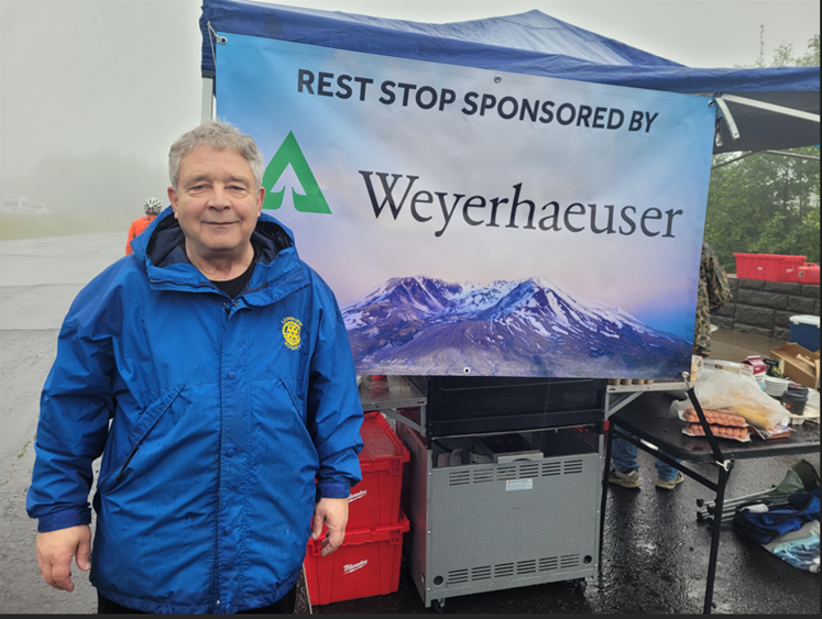 Image of Tim standing at a rest stop sponsored by Weyerhaeuser during the Tour de Bike race. Tim is wearing a blue jacket on a wet day.