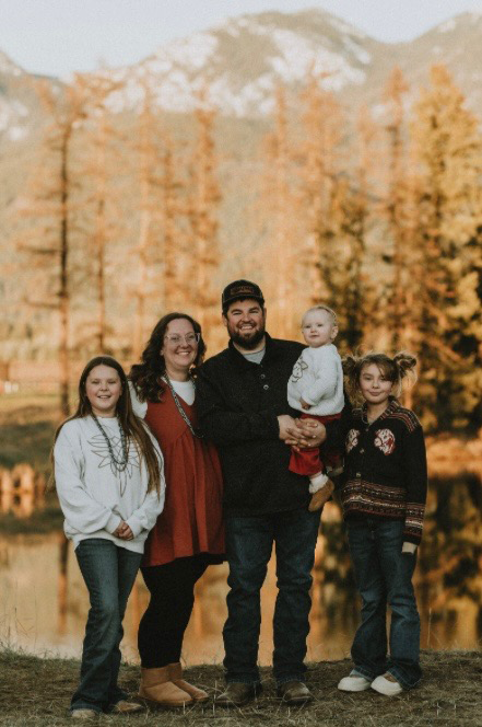 Image of Chance and his family having their photo taken. They are standing in front of a lake with trees and mountains in the background.
