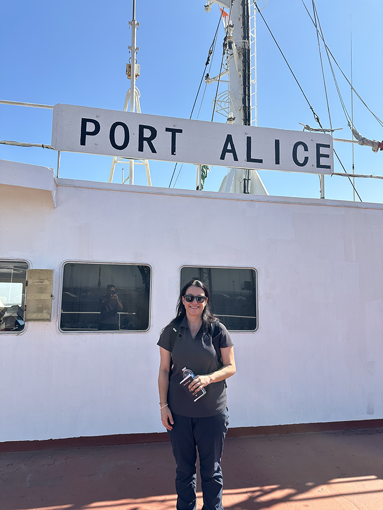 Image of Alice standing in front of a sign for Port Alice as she arrives in India. Kelly is smiling and wearing a brown short-sleeve button-down shirt and dark slacks. Her hair is in a ponytail. She is carrying a water bottle and her passport.