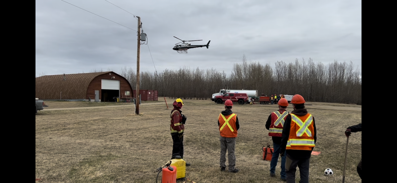 Image of a helicopter in the distance as it prepares to land, with individuals wearing protective gear (vests and hard hats) await its arrival.