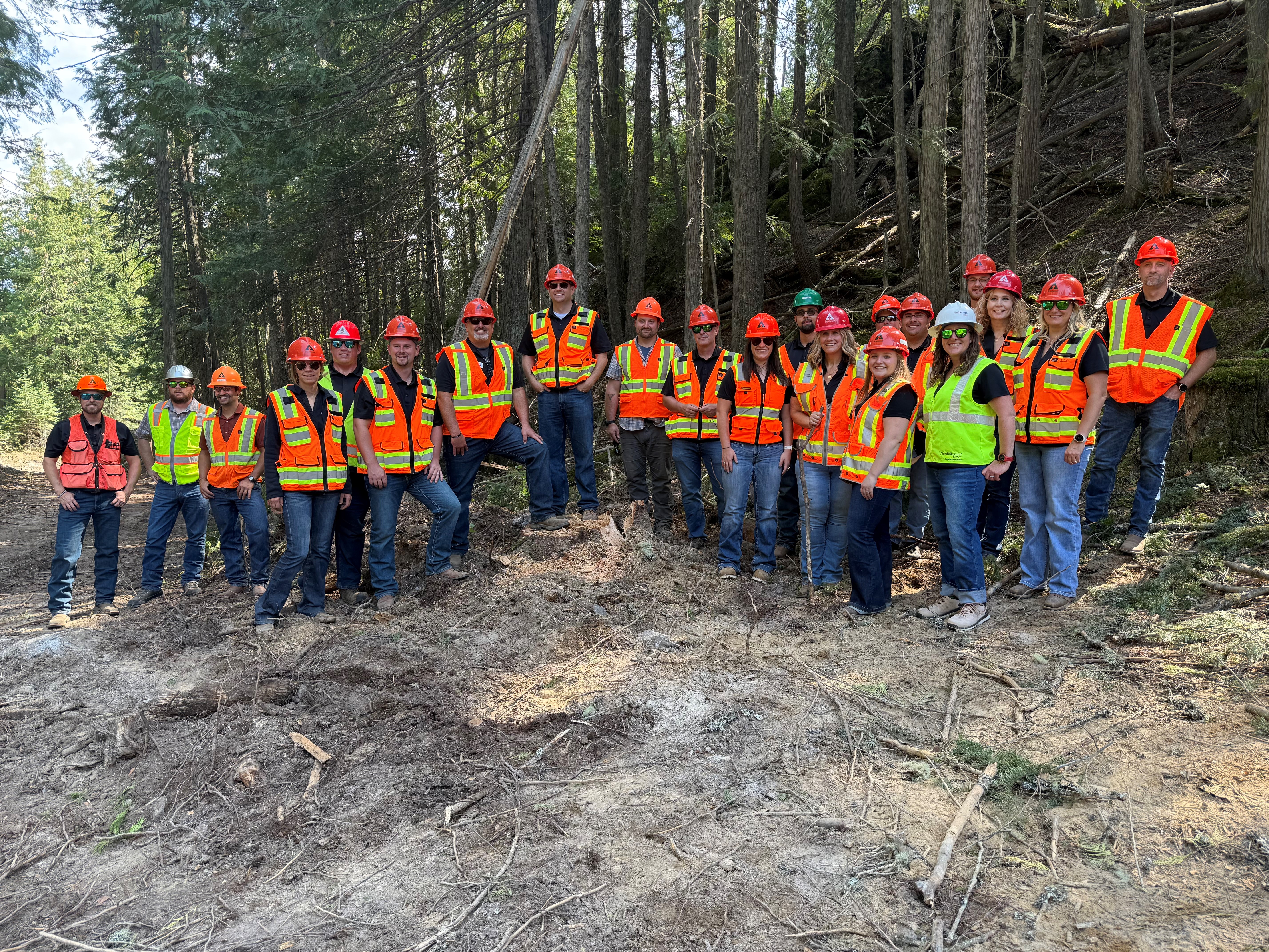 Imagte of the REAL Montana Class 6 participants. The team are at a harvest site with dirt in front of them and trees behind them. All of the participants are wearing protective gear with safety vests and hard hats.