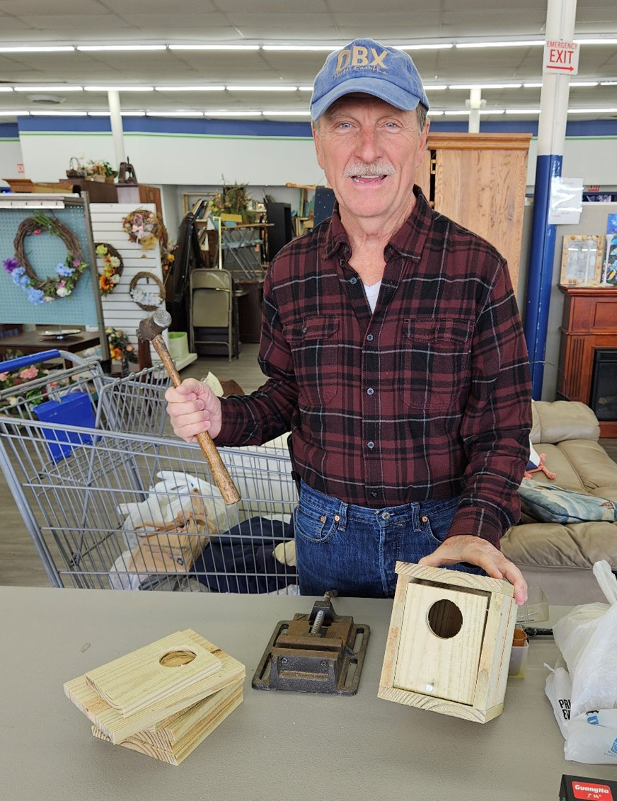 Image of a birdhouse being built from scrap lumber. A man in a red flannel shirt wearing a blue baseball cap holds a birdhouse and a hammer, as other pieces of wood sit in front of him.