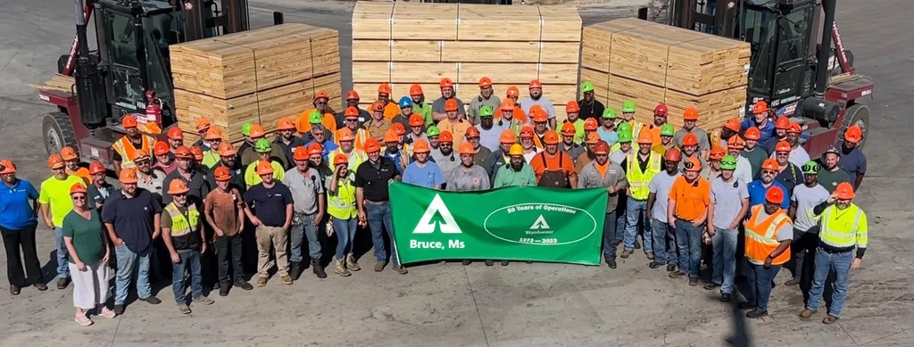 Image of the Bruce team gathered for a photo. Dozens of team members are standing in front of stacks of wood while wearing personal protective gear and holding a green banner.