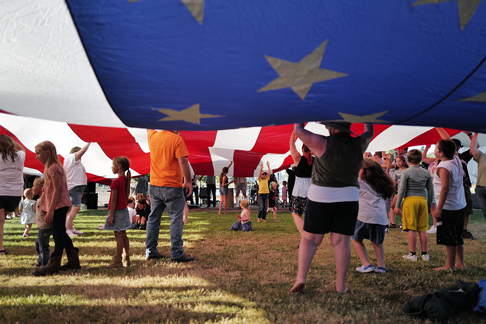 Image of a young child sitting on the ground under a massive flag of the United States of America.