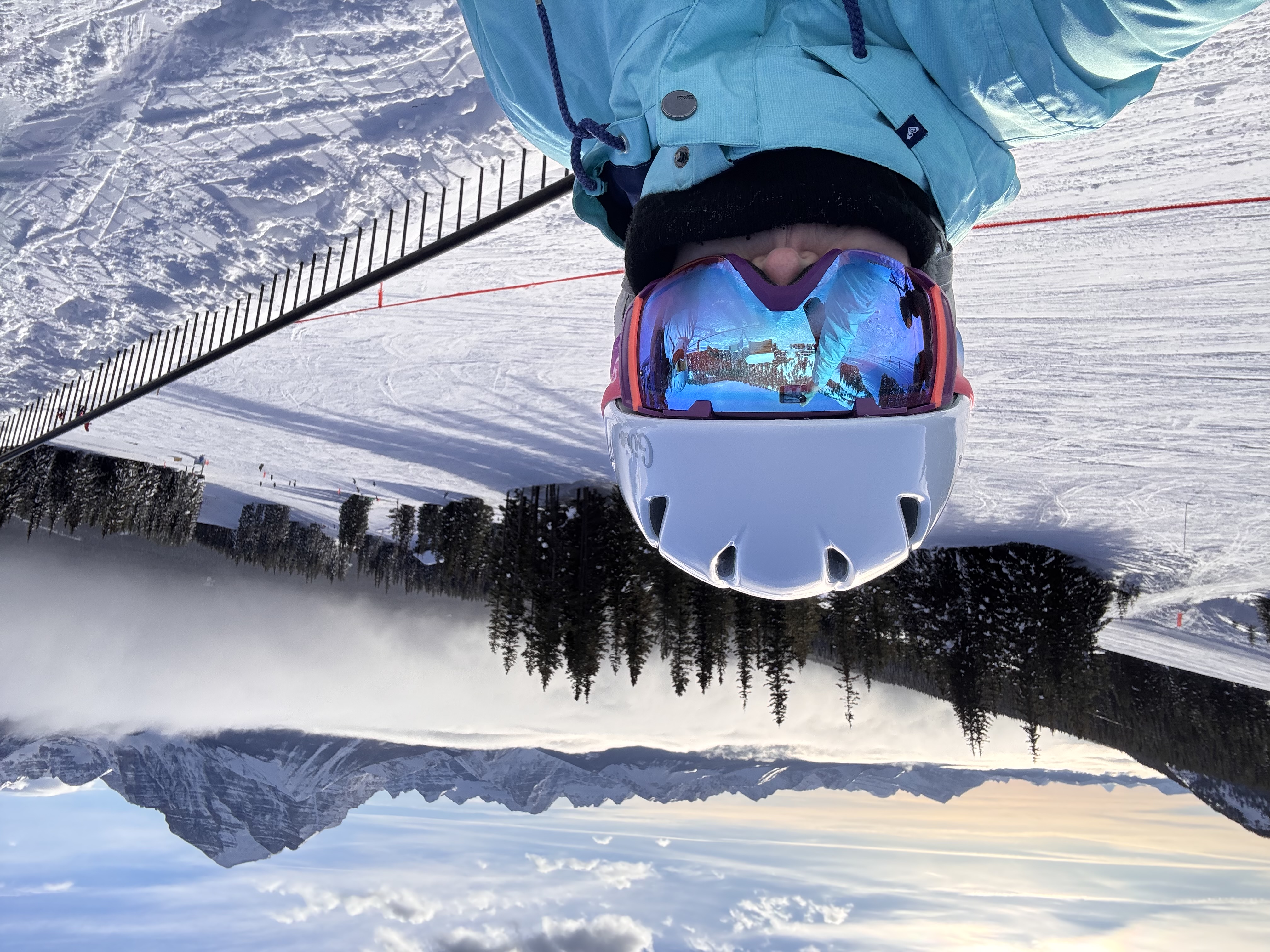 Image of Kerri skiing. She is wearing a helmet and large goggles and her back is to a snow-covered hill. Clouds can be seen in the background.