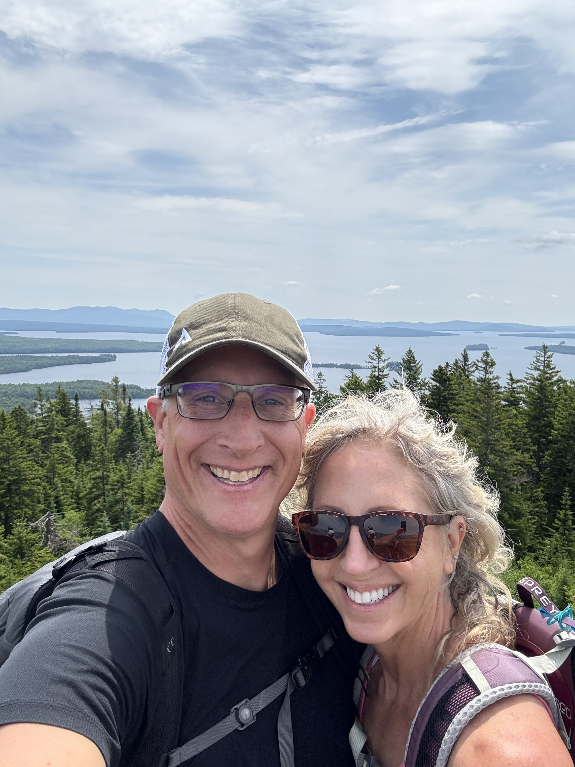 Image of Michelle with her husband Erik. They are each wearing backpacks. Michelle wears sunglasses while Erik wears glasses and a cap as they take a selfy in front of the lake with fir tress in between the lake and Michelle and her husband.