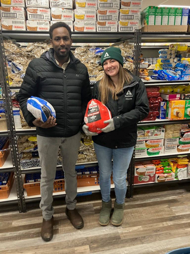 Image of Abdul and his colleague Jasmine Hagen holding turkeys in the food bank. Adul is standing to the left and Jasmine to the right. They are each wearing coats. Abdul's turkey is in a white and blue package, while Jasmine's is in a red package. Jasmine is also wearing a green Weyerhaeuser knit hat.
