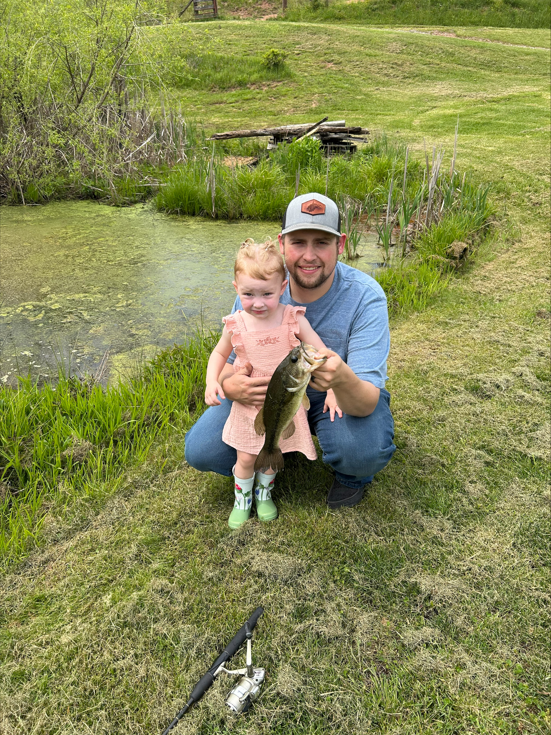 Image of Nathan with his daughter Harper holding a fresh fish that they caught. Nathan is wearing blue jeans and a blue shirt with a baseball cap. Harper is wearing a pink dress with frilly straps and a pair of rainboots.