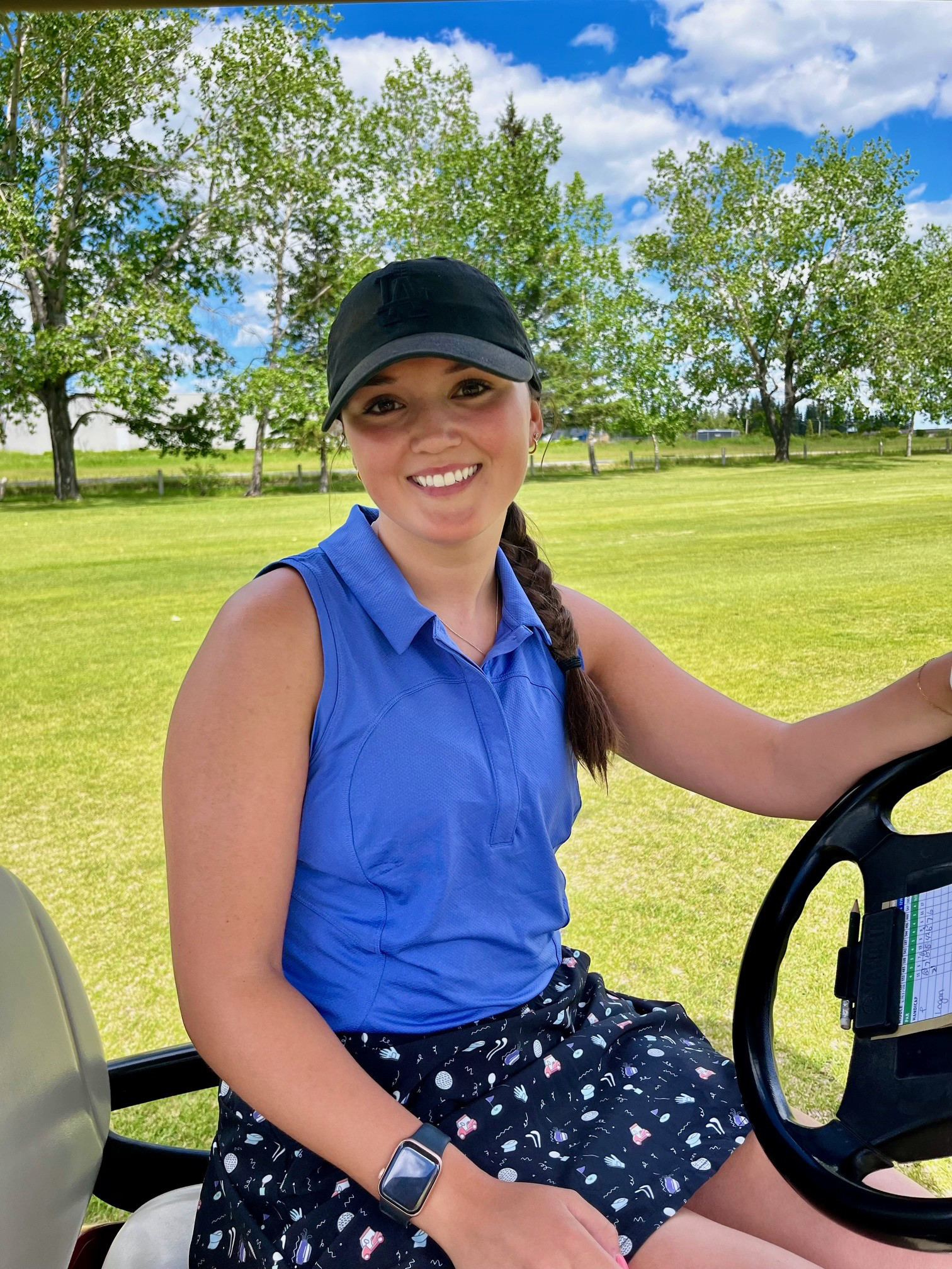 Image of Logan sitting in a golf cart. She is wearing a black hat with a blue sleeveless shirt and dark skirt with patterns. In the background are a group of trees and beautiful grass.