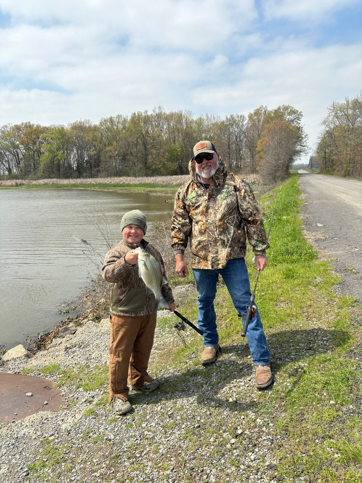 Image of Joe and John Henry at the side of a lake. Each are wearing camouflage jackets and John Henry is holding a white bass.