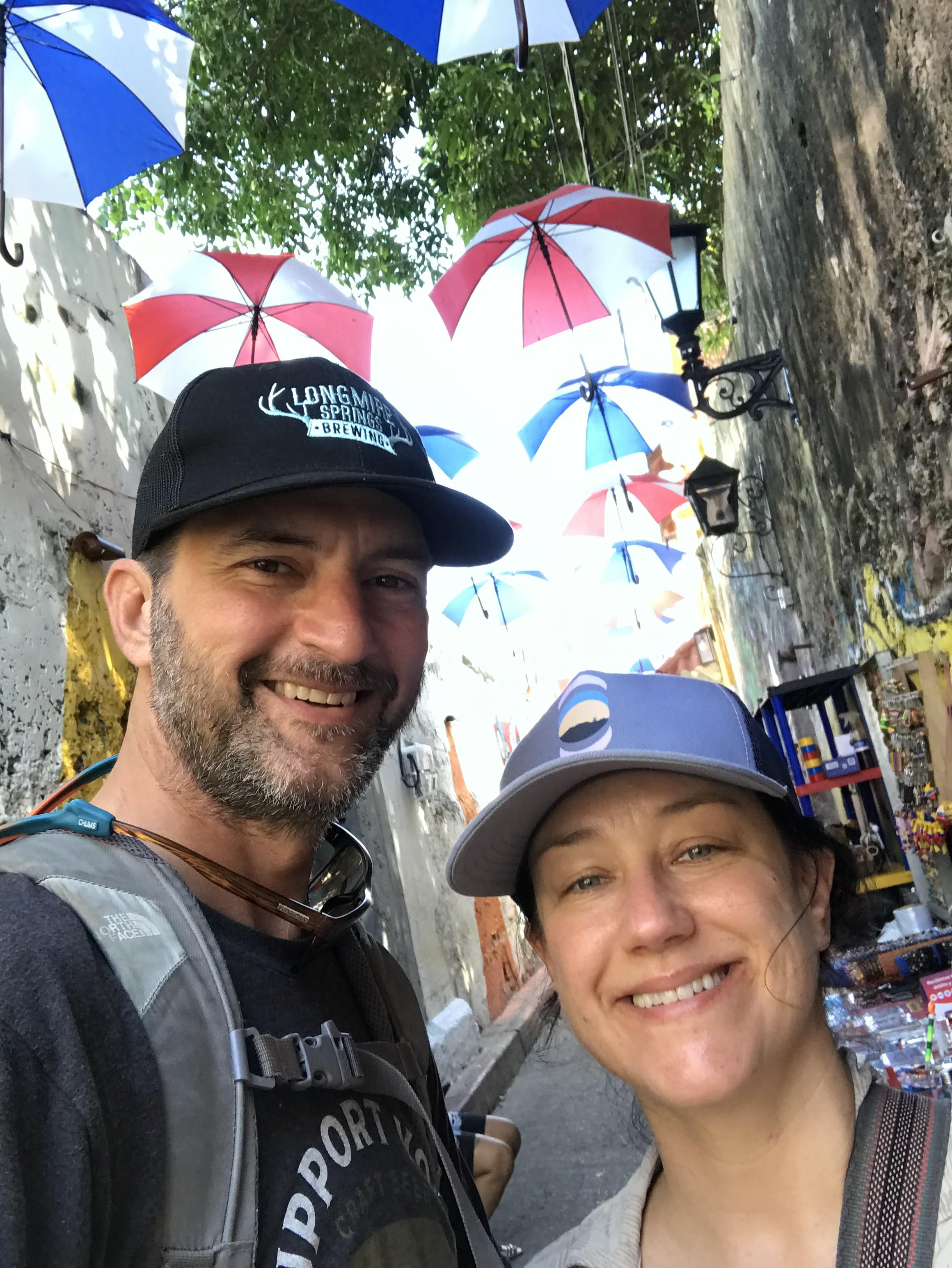 Kelly and her husband in Cartagena, Colombia. She is wearing a blue baseball hat and her husband is wearing a black baseball cap. In the background umbrellas line the second floors of the street.