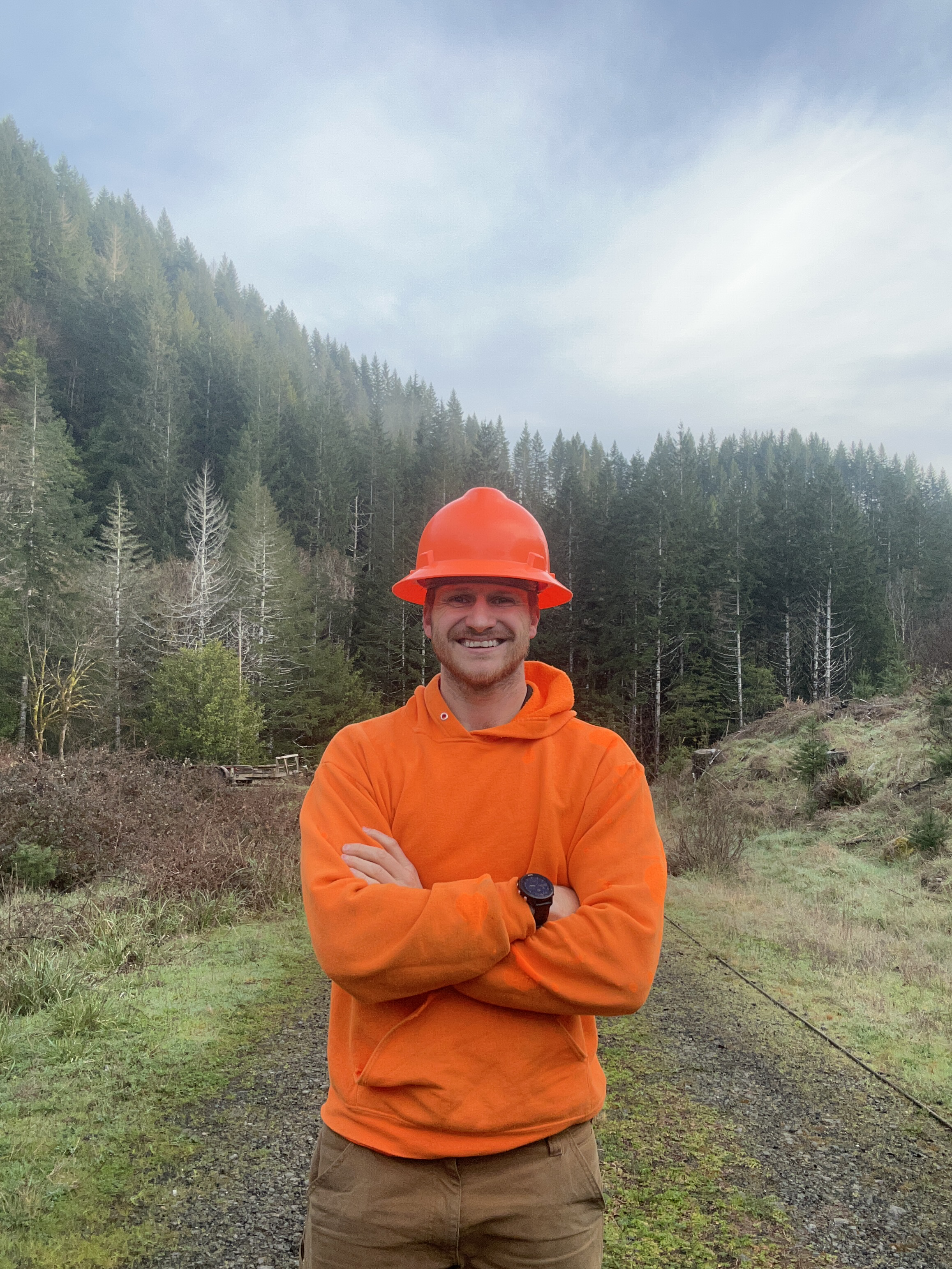 Image of Ky Smith wearing an orange hard hat and orange hoodie as he's pictured with the work being done behind him.