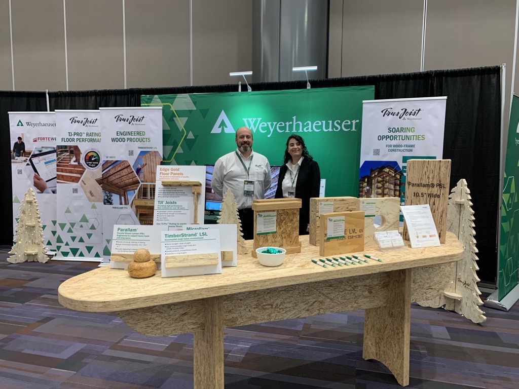Image of Neda and Steve McManus standing in a Weyerhaeuser booth at a convention. A large, green Weyerhaueser sign is directly behind Neda and Steve, and displays of products can be seen all around.