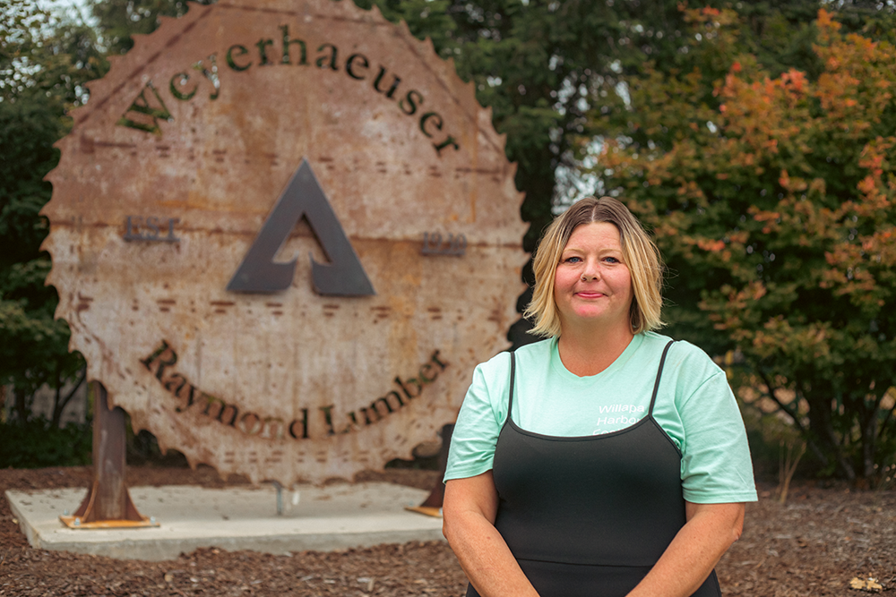 A woman stands in front of a carving of a saw blade made out of wood with Weyerhaeuser's Raymond facilitiy's name on it.