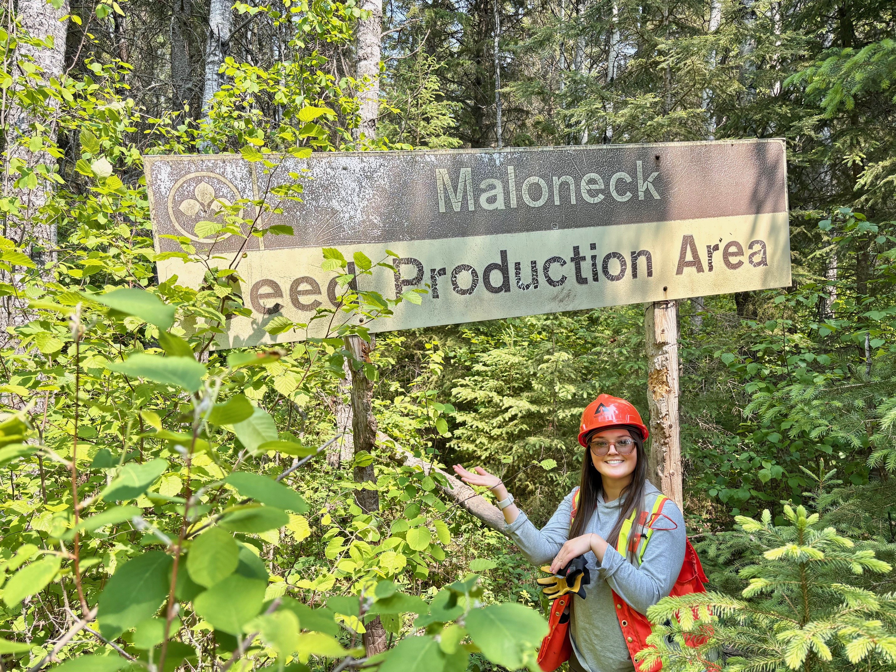 Logan stands in front of a sign that reads "Maloneck Seed Production Area." She is wearing an orange helmet and orange safety vest.