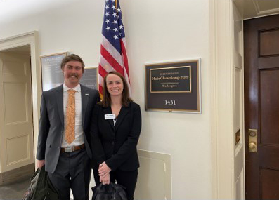 Image of Colin Towne and colleague Megan Shultz on a trip to Washington, D.C. They are each wearing suits and are standing outside an office and in front of an American flag.