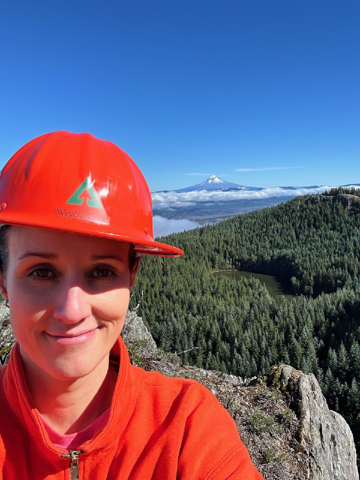 Image of Jill, wearing an orange Weyerhaeuser hard hat and orange fleece, while taking a selfie with Mount Hood in the background of the image. Fields of trees can be seen between Jill and Mount Hood.