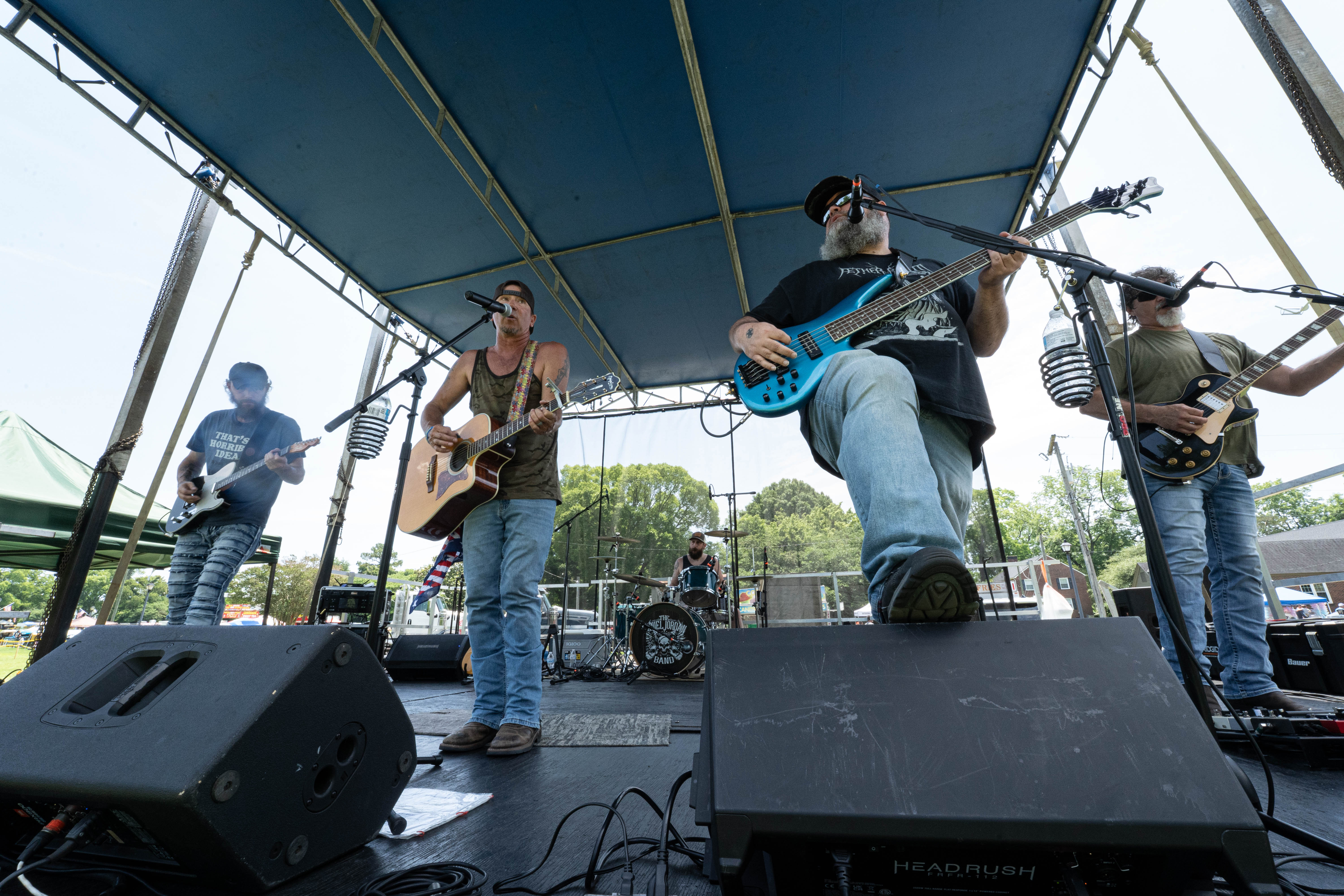 Image of perrformers on stage at the festival. A band plays with the bass player placing his foot on the amplifier in front of him as three guitar players are along side him, and a drummer in the background.