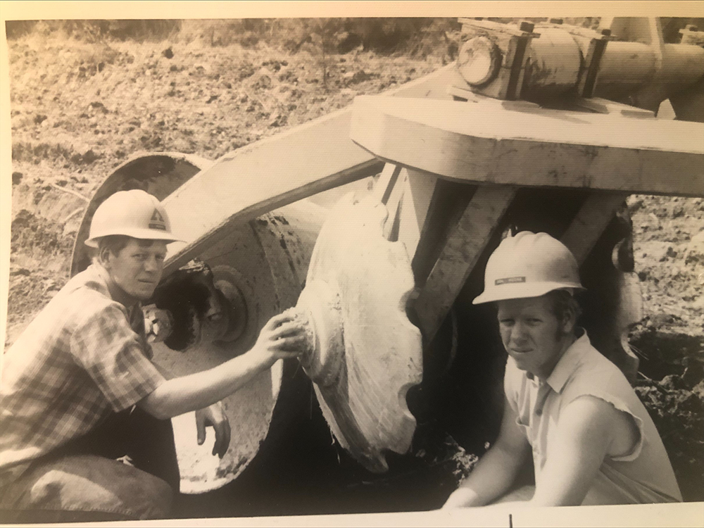 Black and white image of Don and his twin brother Lon as they inspect a bedding plow. Don is on the right side of the image and is wearing a shirt with cut-off sleeves, while Lon, on the left, wears a flannel shirt. Both are wearing hardhats and are crouched down while inspecting a large piece of machinery.