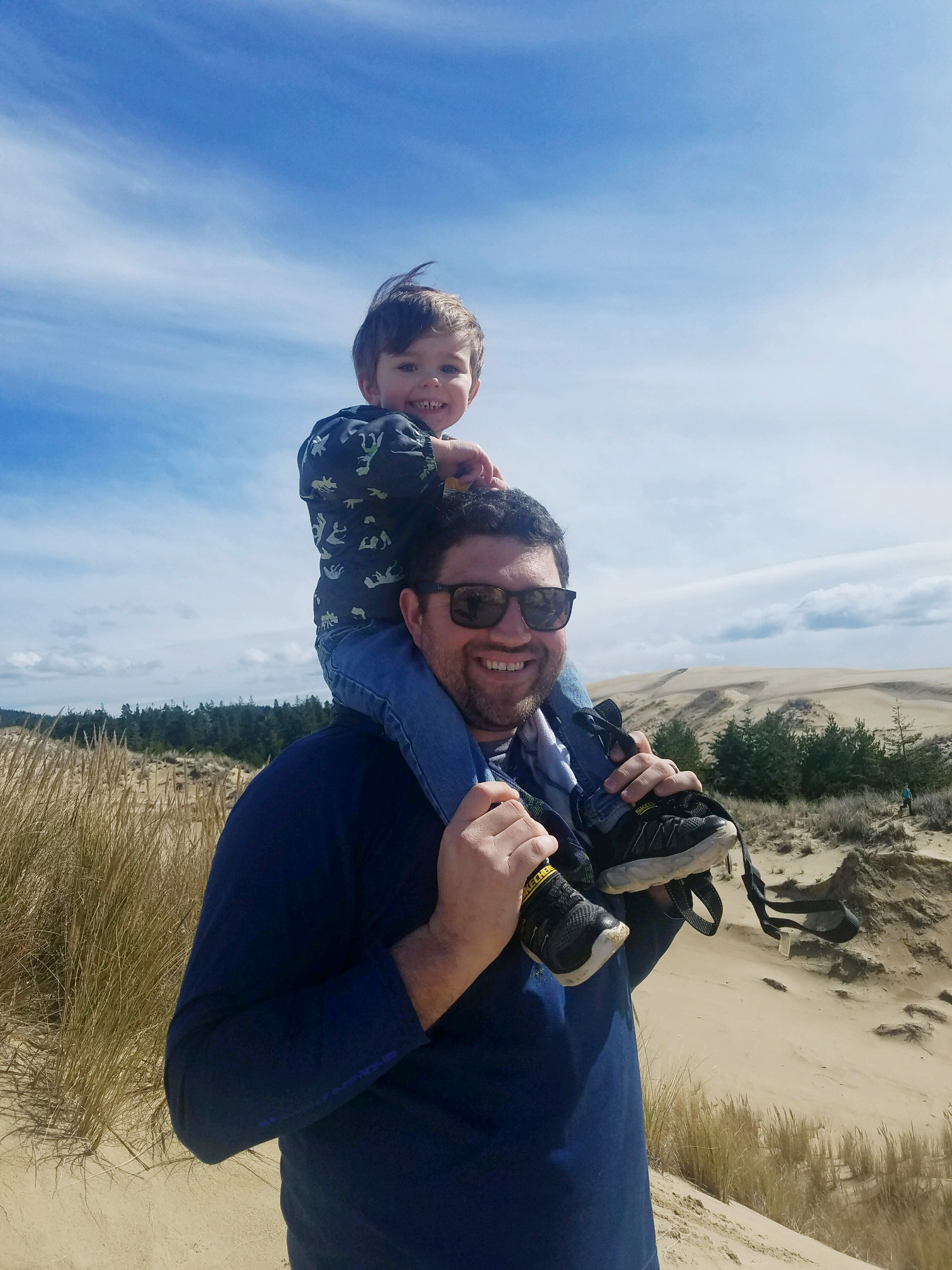 Image of Seth and Nolan exploring the sand dunes on the Oregon coast. Nolan is sitting atop Seth's shoulders as Seth holds onto Nolan's ankles. Both are smiling. Seth is wearing sunglasses.