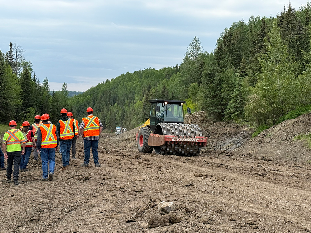 Image of a group of employees wearing safety vests and hard hats and walking past a vibratory packer as it works. 