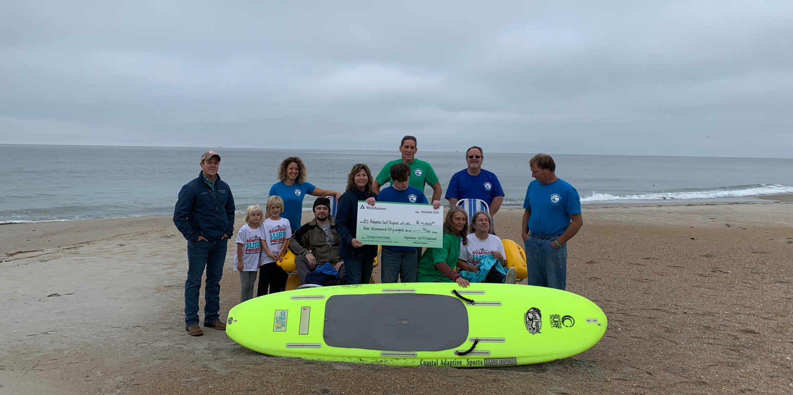 Image of Lori presenting a Giving Fund check to the Adaptive Surf Project. Lori and a number of people pose with the check as a surfboard is in front of them and the ocean is behind them.