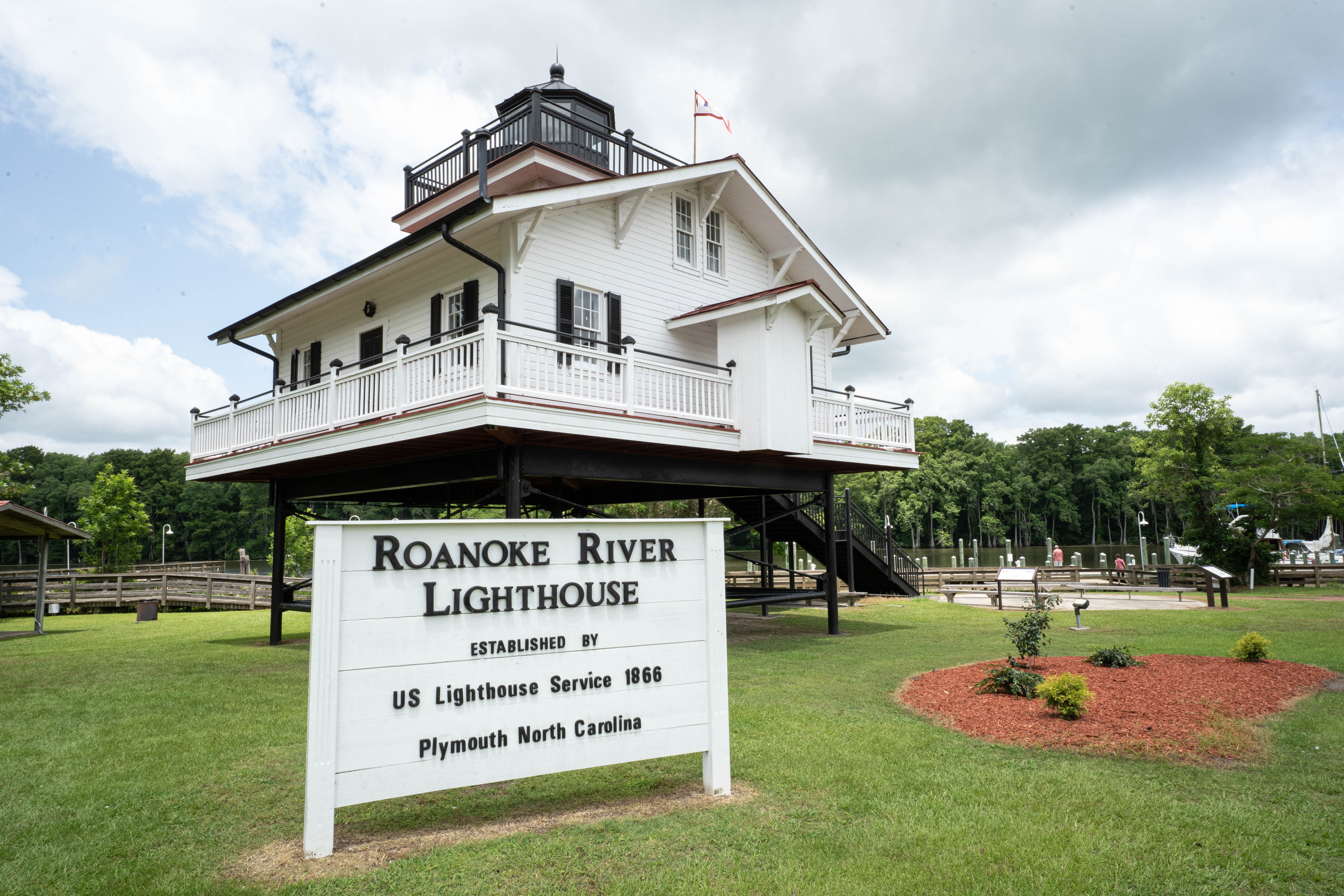 Image of the Roanoke River Lighthouse. The lighthouse stands on black stilts above the ground, The lighthouse itself is white with a black turret atop from whiich a viewer can see 360 degrees.