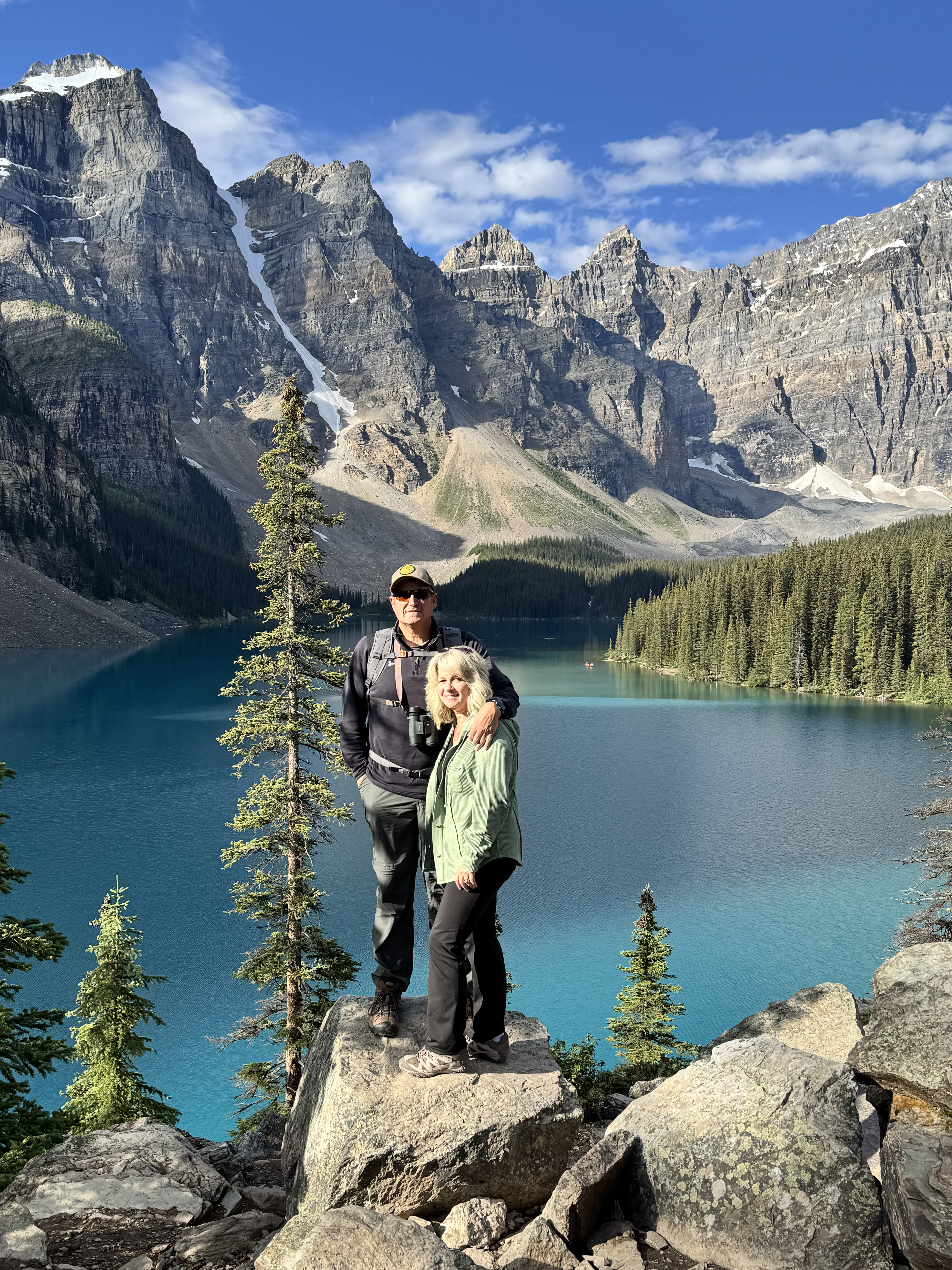Tim and his wife Cindy stand in front of Moraine Lake in Banff, Alberta. There is a single tree standing behind them as they stand on rocks overlooking the lake with mountains rising in the background.