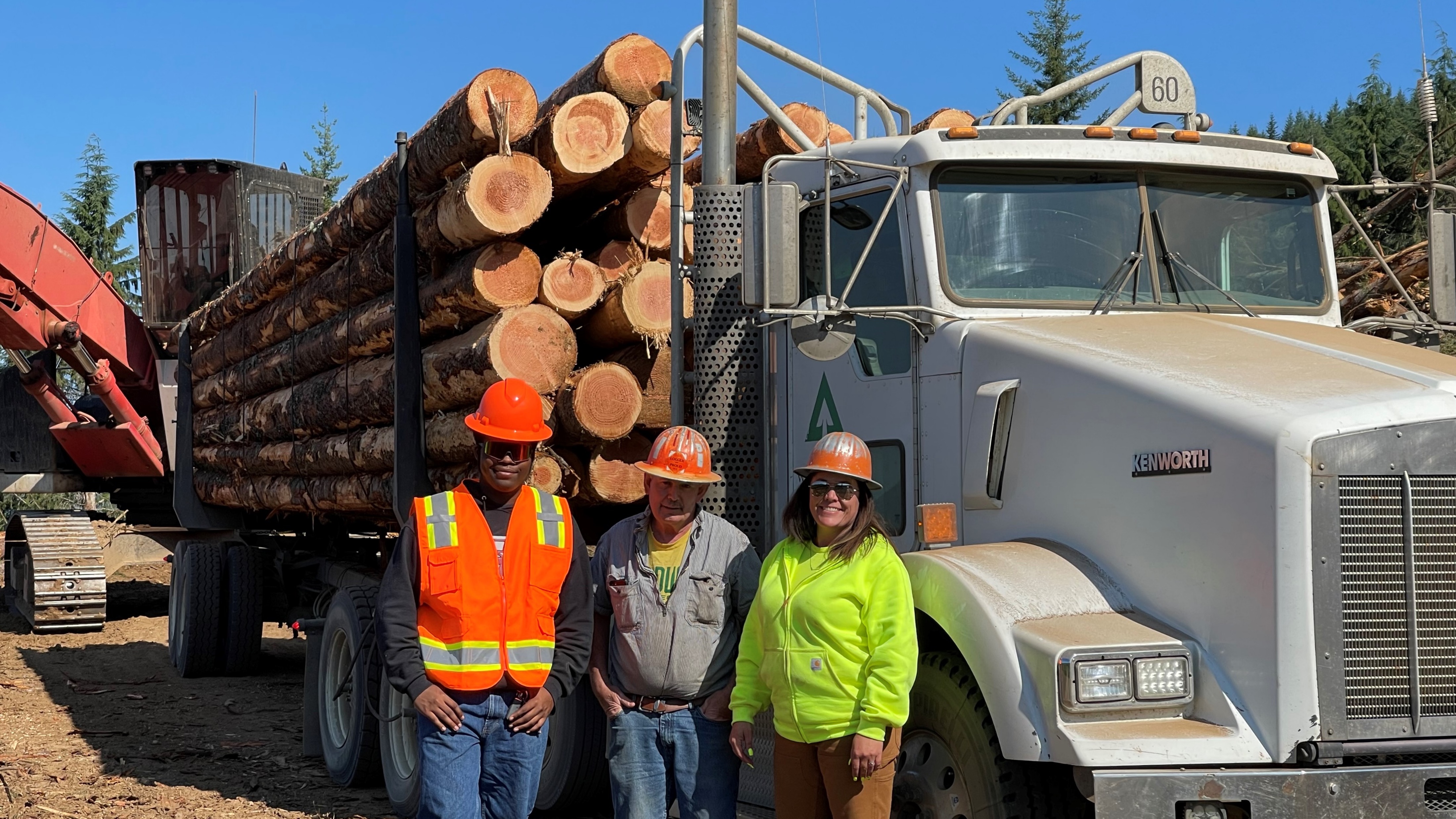 Image of Danny Simili, shovel loader Rick Phillips and Leesha Carson in front of a loaded log truck.