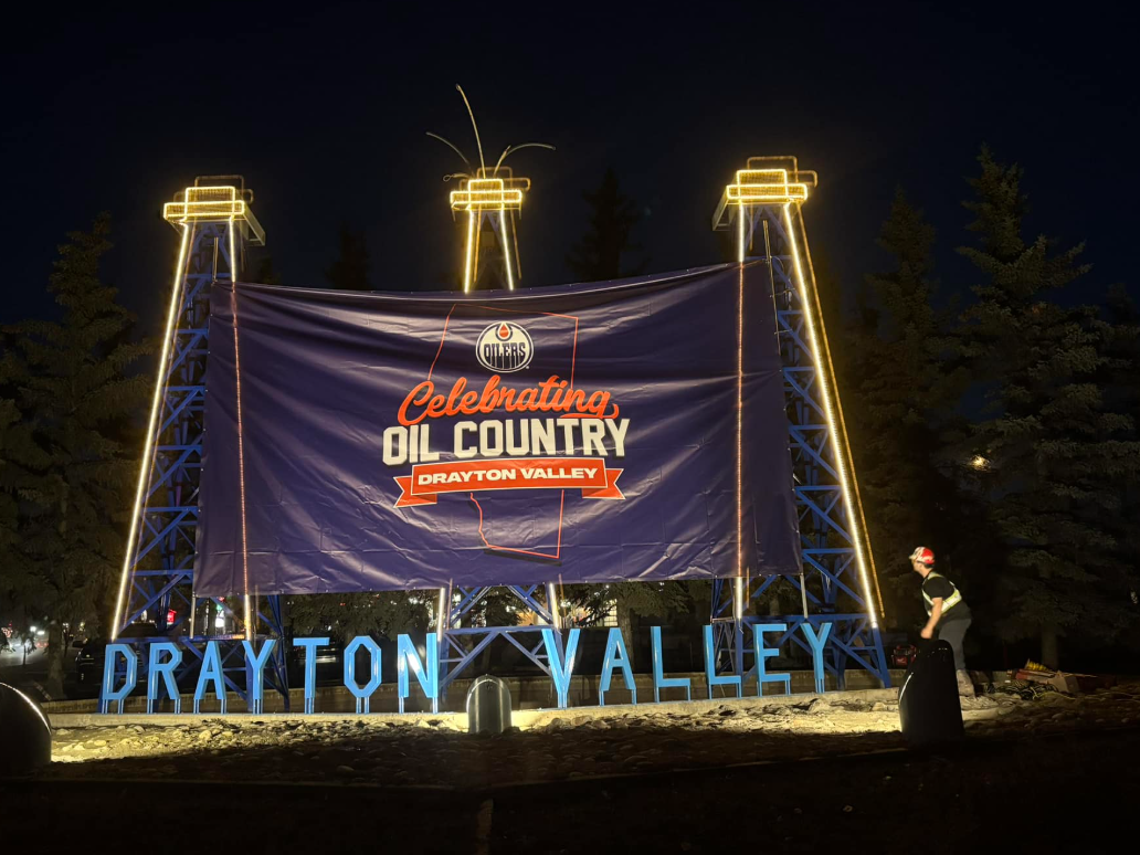 Image of a new addition to the Drayton Valley sign, a banner that says "Celebrating Oil Country" strung among three fake oil derricks.