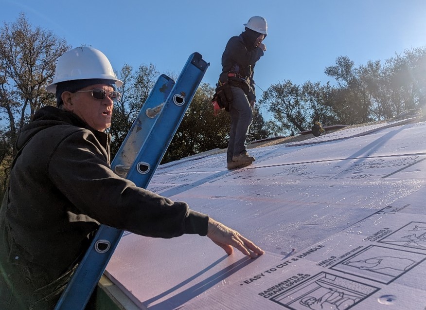 Image of volunteers working on a home in Ogden, Kansas. One man wearing a hard hat stands on a roof while another, also wearing a hard hat, is on a ladder.