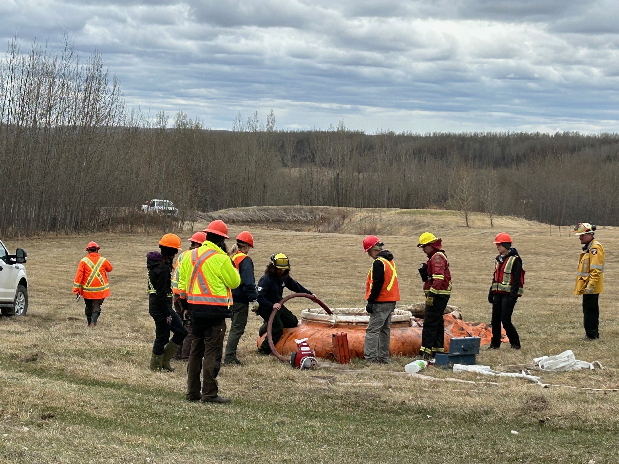 Image of numerous employees wearing protective gear of bright jackets and hard hats training to fill a portable water bladder. They are in a field with barren trees in the background.