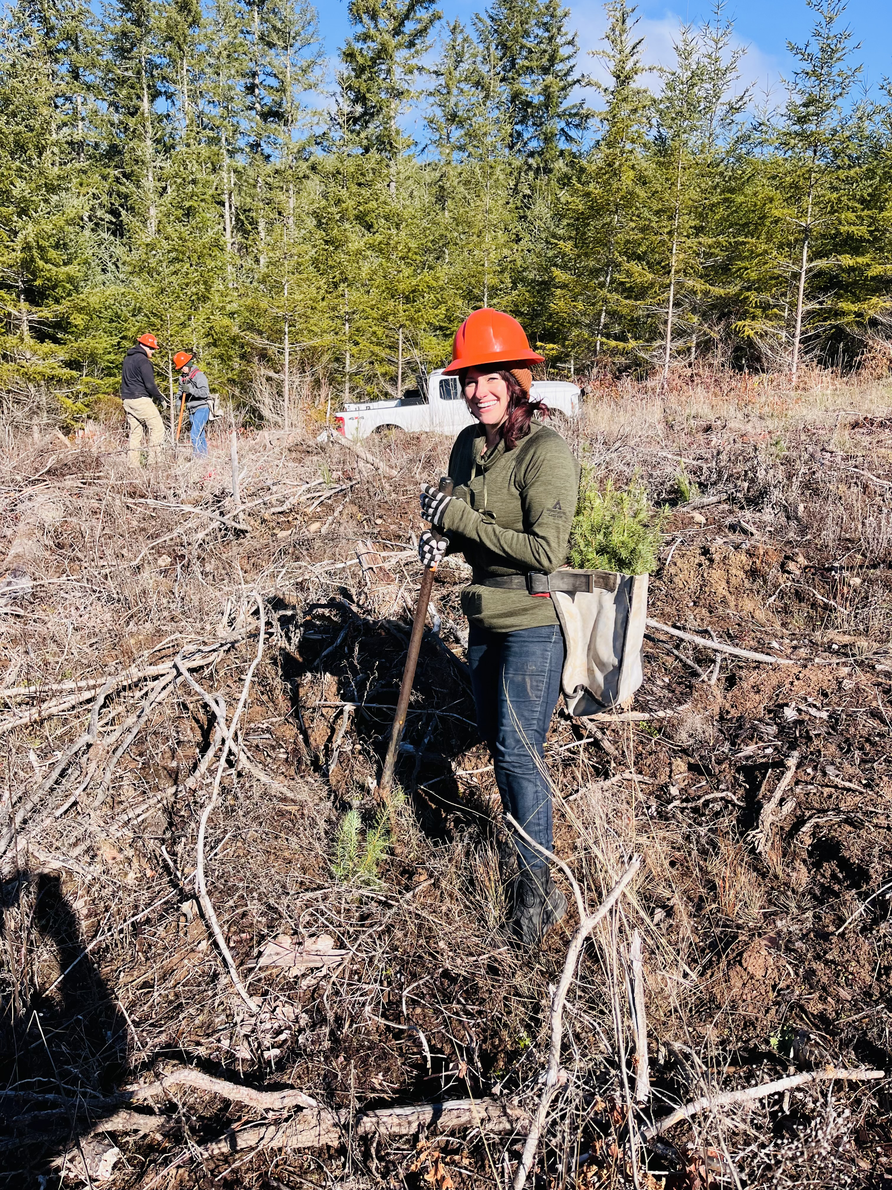 Image of Land Use Manager Lyndcy Patrick in the field. She is wearing an orange hardhat, a green jacket and jeans. She is in a clearing in a forest and is holding a shovel handle.