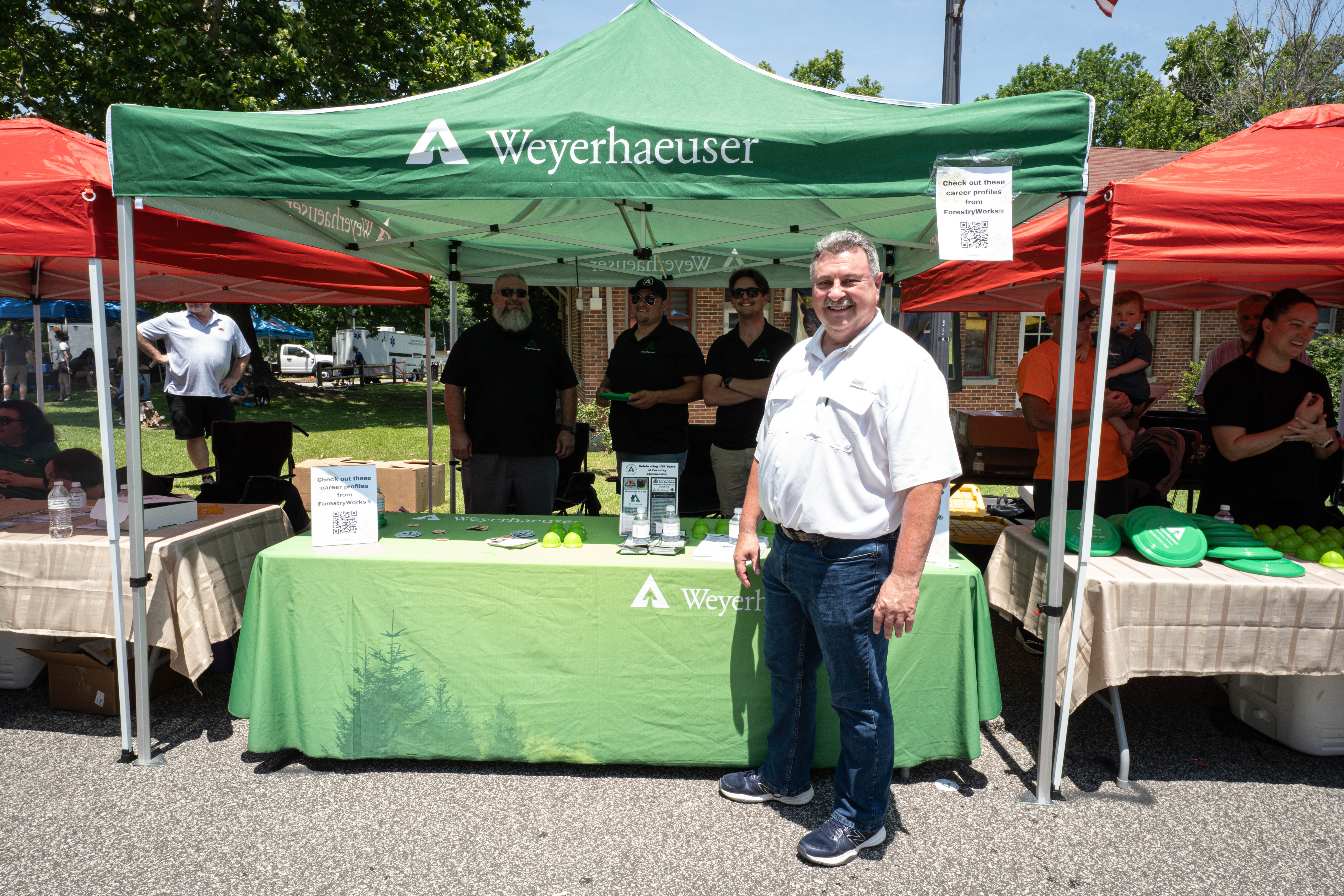 Image of the Weyerhaeuser booth from the Black Bear Festival. Plymouth Mayor Brian Roth stands in front of the booth, which is green with a green table in front.