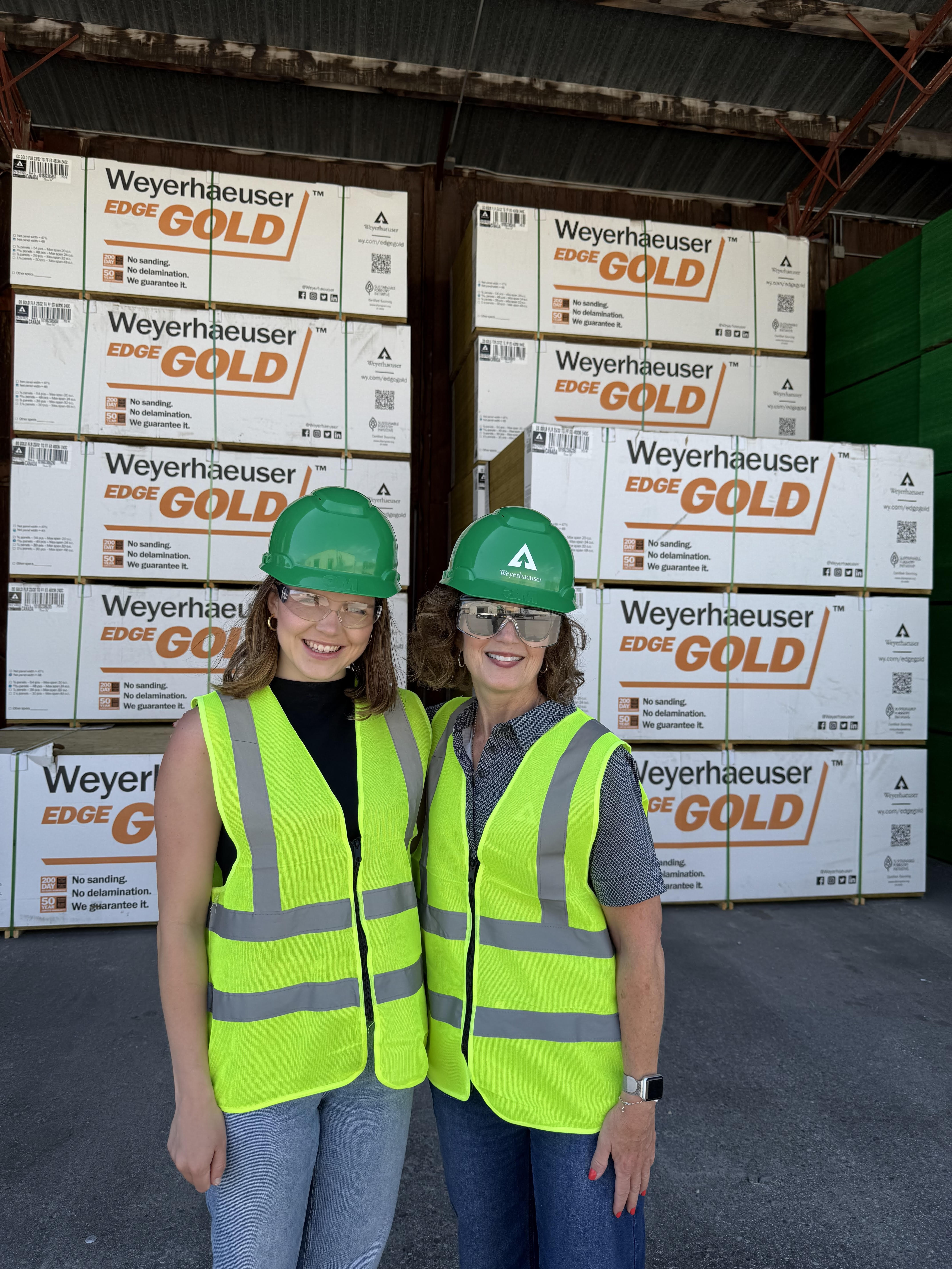 Image of Ashely and a colleage wearing green hardhats and yellow safety vests while standing in front of a massive stack of lumber.