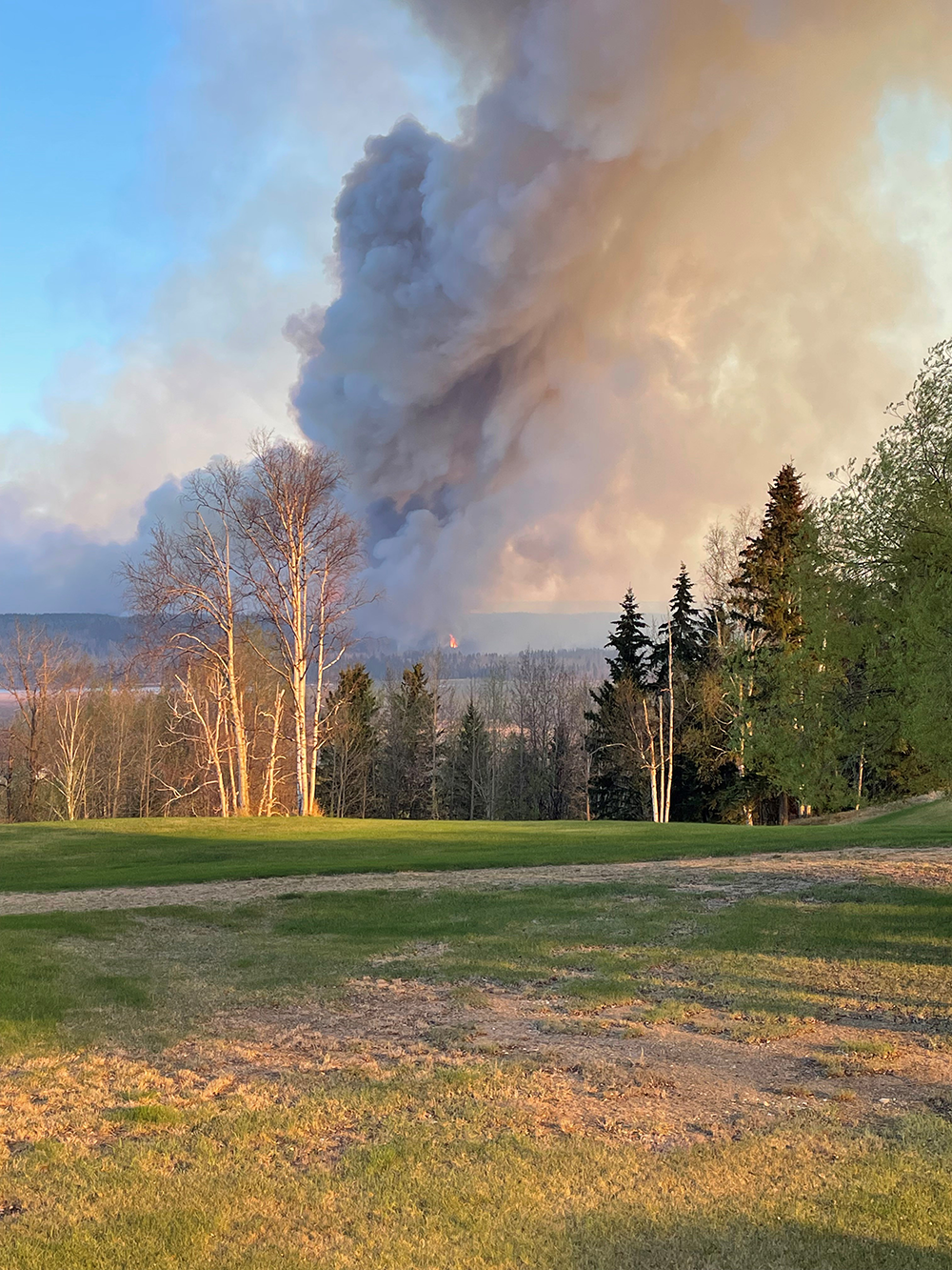 Image of a wildfire approaching a populated area. Large amounts of black and gray smoke rise in the background of the image with trees and fields in the foreground.