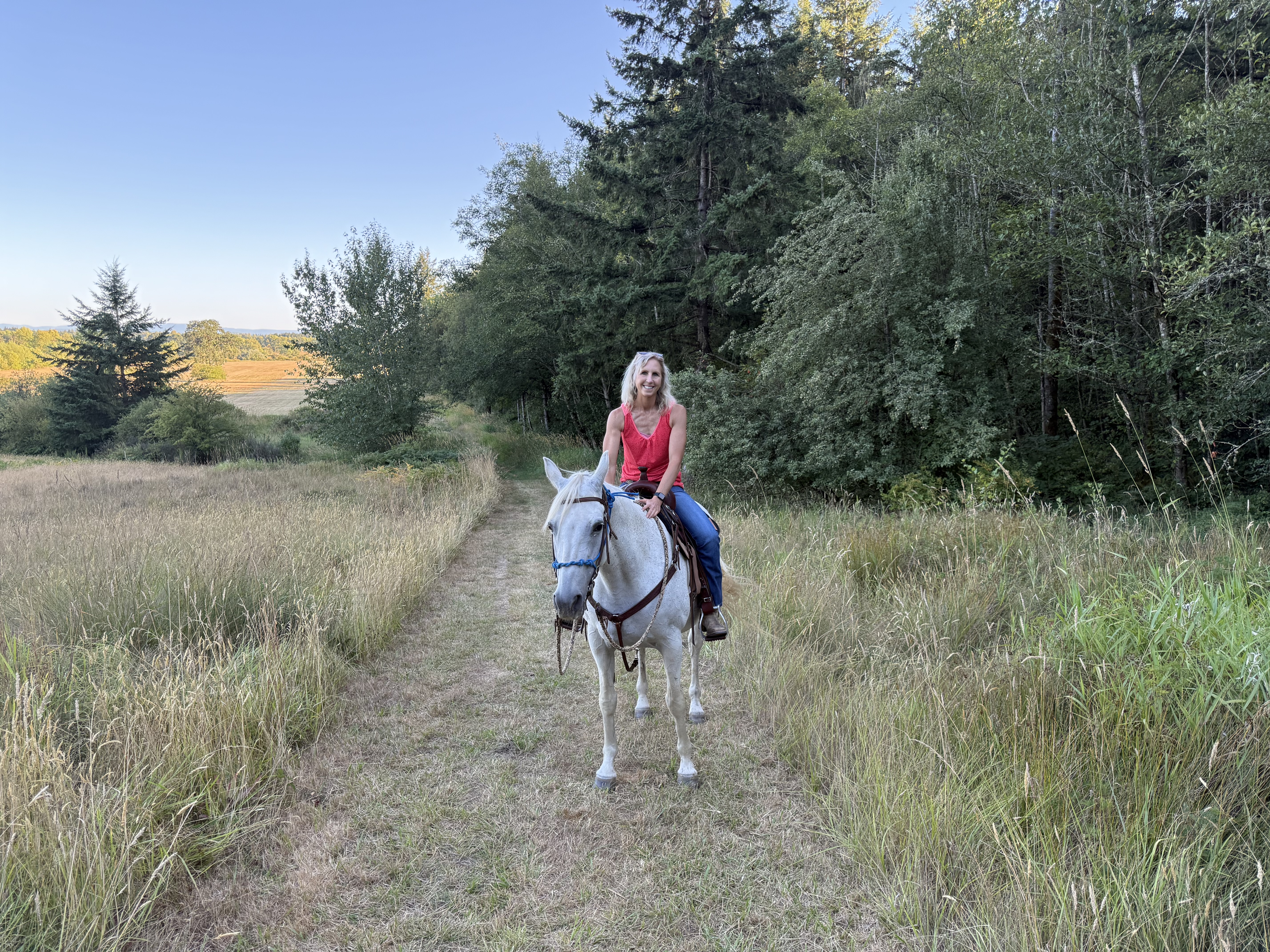 Image of Michelle riding her horse Lexi. Lexi is a white horse and Michelle is wearing a red blouse They are surrounded by trees and grass on Lexi's left.