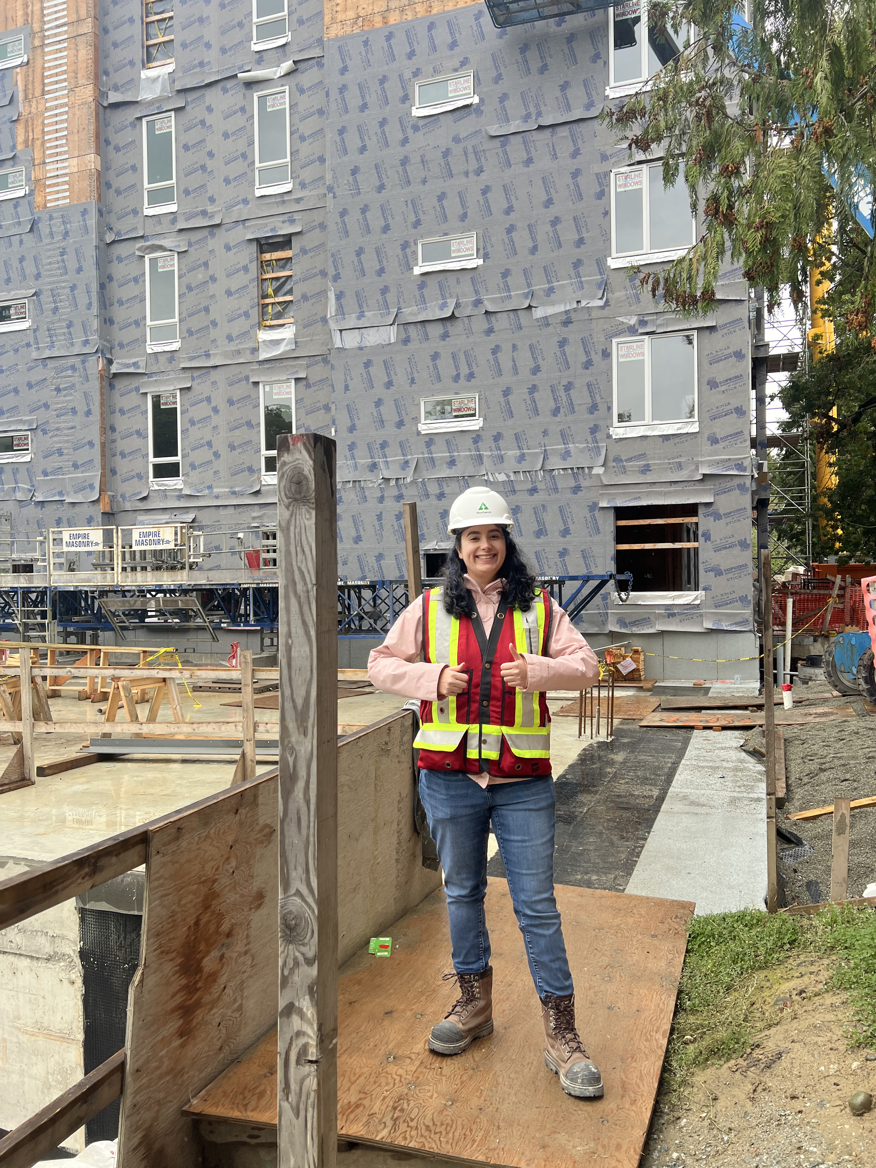 Image of Neda on a job site. She is wearing PPE, including a safety vest and hard hat, and is holding two thumbs up.