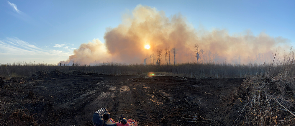 Image showing a fire in the background of the image, with huge amounts of gray smoke rising through the skyline. Burned fields and charred trees can be seen in the foreground.