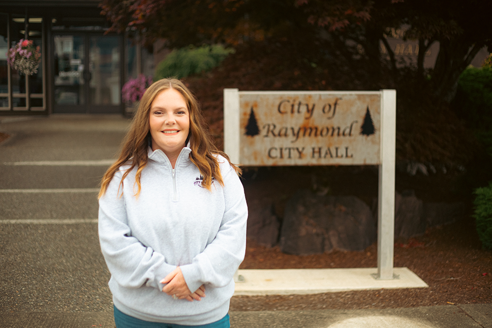 Woman in a white hoodie stands in front of Raymond City Hall.