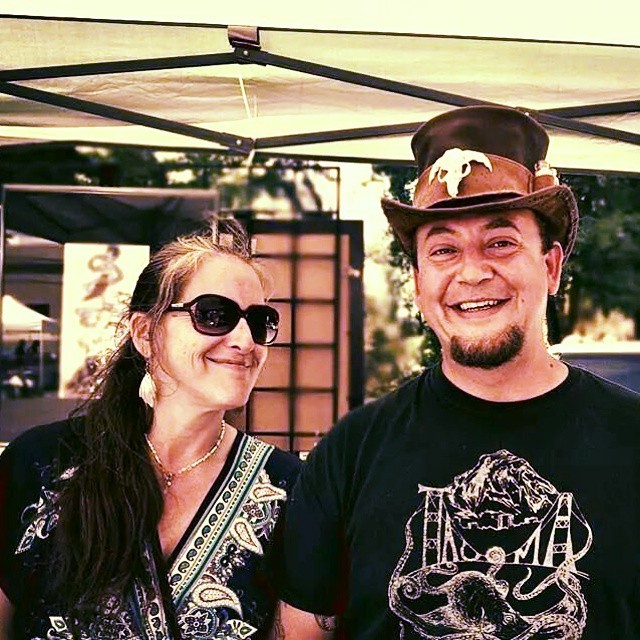 Bob and his wife Carrie, right, are pictured at an art fair. Bob, on the left, is wearing a stempunk top hat and a black t-shirt with Carrie's design on it in white ink. Carrie is wearing black sunglasses and has long hair and is looking at Bob, smiling.