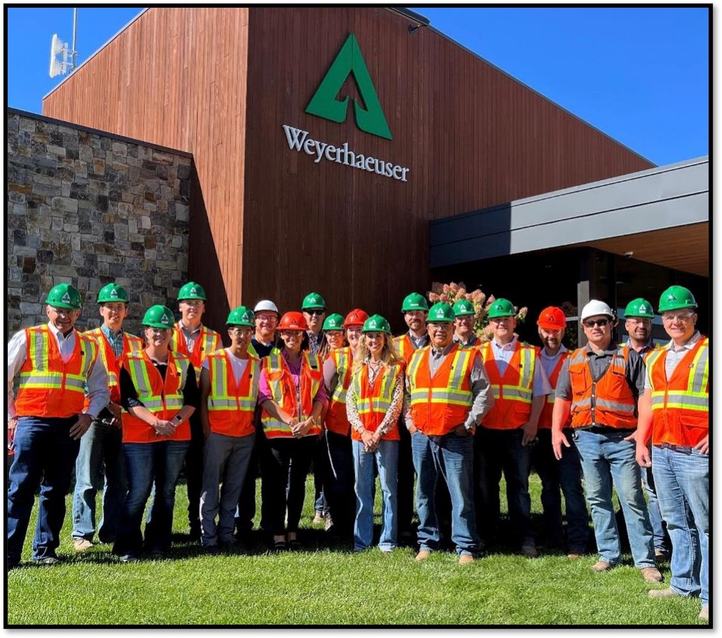 The REAL Montana class poses in front of a Weyerhaeuser building. All members of the team are wearing PPE, including safety vests and hard hats.